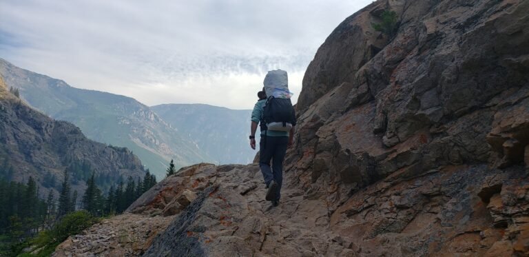 A backpacker carrying an ultralight pack walks along a rocky mountain ridge with a broad valley and distant peaks in the background.