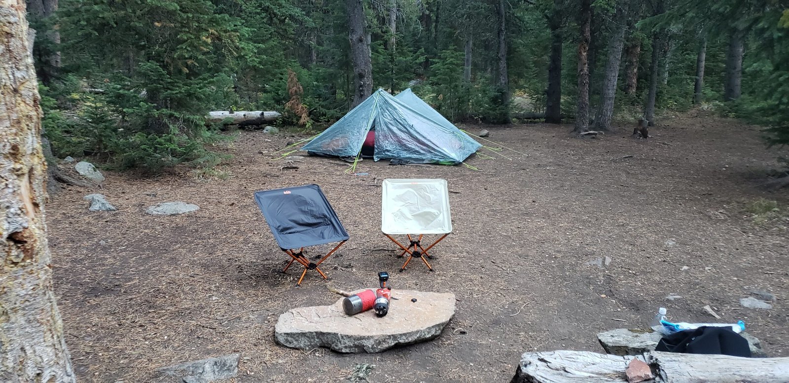 A small ultralight tent and two camp chairs at a quiet wooded campsite, with simple gear arranged on a flat rock in the foreground.